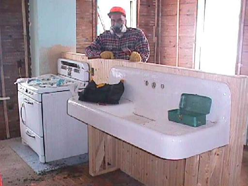 Brian stands behind the half-wall which supports the white gas range and five-foot wide sink with double drainboards