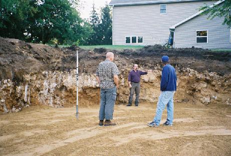 Ken addressing Brian and Travis in basement hole with backdrop of topsoil, clay and bedrock