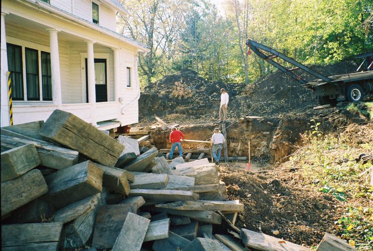 wood beam pile in 
	foreground, towtruck winch in background, all around excavated hole