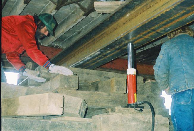 workers on crib of wood beams supporting hydraulic jack for lifting metal I-beam
